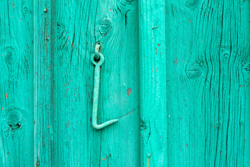 Close-up of a vintage metal hook on a turquoise painted wooden wall. The weathered paint and textured timber create a rustic, distressed look, ideal for farmhouse design or industrial backgrounds