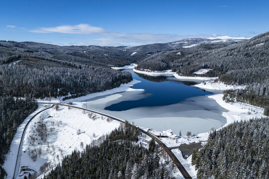 Aerial view of a serene, dark blue lake nestled amidst snow-laden forests, punctuated by a stark white blanket and a winding road, Soboth, Styria, Austria.