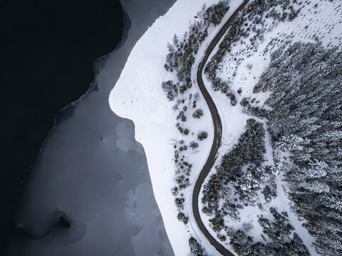 Aerial view of a sinuous asphalt road winds along the snowy banks of Stausee Soboth lake, bordered by a dense forest of snow-laden trees, Soboth, Styria, Austria.