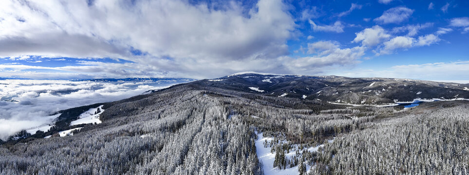 Aerial view of a snow-laden forest stretches across rolling hills towards a distant lake under a sky of mixed clouds and bright blue, Soboth, Styria, Austria.
