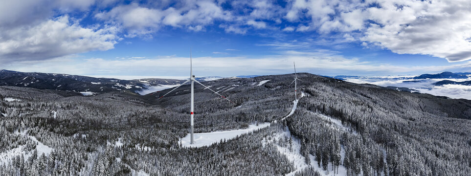 Aerial view of wind turbines stand tall amidst a snow-dusted forest, contrasting against the crisp blue sky and distant, cloud-veiled mountains, Soboth, Styria, Austria.