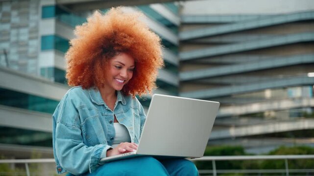 Smiling young latin woman with curly hair using laptop pc computer outdoors near modern building. Student or freelancer typing, working remotely or studying online sitting at university campus