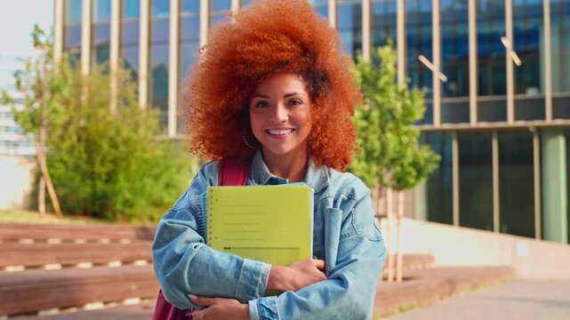 Portrait of African American girl with curly afro hairstyle standing with backpack, holds exercise books in university campus. Brazilian redhead student looking at camera ready for educational course