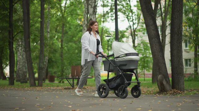 Mother pushing stroller confidently forward. Woman with child on walk through leafy urban setting with intention. Mother and child navigating bustling park avenue promptly with purpose and composure