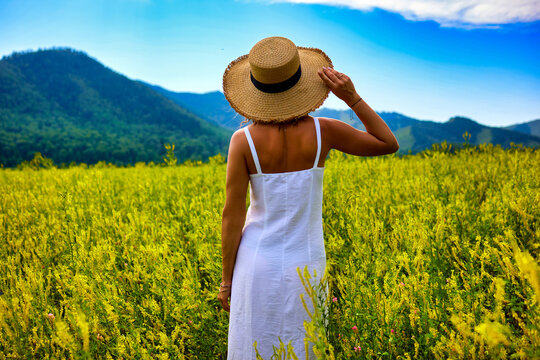 A radiant woman in a flowing white linen dress and elegant straw hat stands surrounded by vibrant mustard blooms, with breathtaking mountains and a bright blue sky creating a picturesque background.