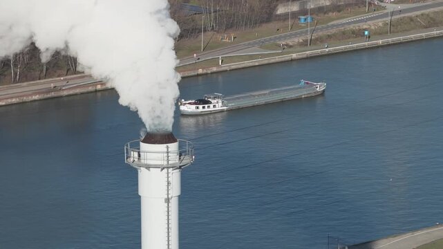 25 february 2026, Genk, Belgium. Aerial Drone Shot of Smoking Industrial Chimney with Cargo Barge Passing on Canal, Air Pollution, CO2 Emissions and Inland Shipping Concept. Inudstrial smokestack, hea