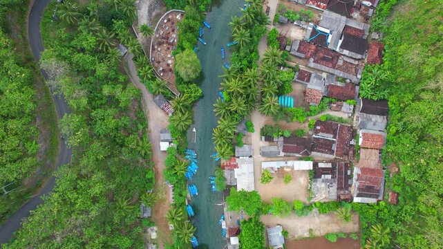 Aerial drone footage from above of a hidden local village in the middle of a vast forest and mountains, with Maron river and blue touristic wooden boats, in Pacitan regency, Java island, Indonesia