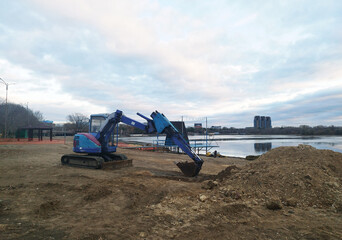 Fototapeta premium A small blue and purple excavator sits on muddy, brown ground next to a calm body of water against a wide, cloudy sky at dusk or on an overcast day.