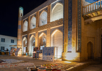 Obraz premium Khiva, Khorezm Province, Uzbekistan - May 30, 2025: Night View of Illuminated Historic Architecture and Traditional Suzani Textiles Market in the Ancient City of Khiva