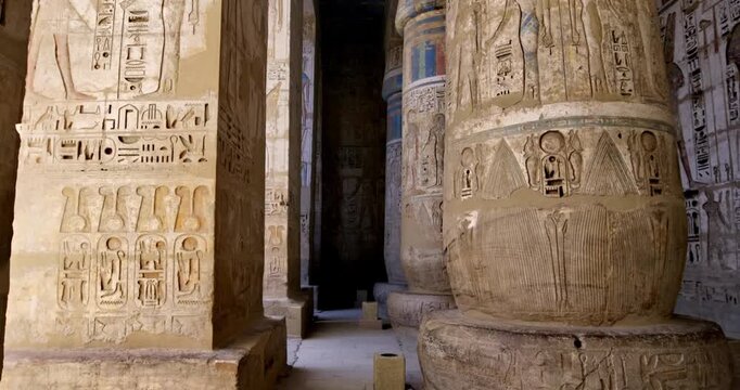Medinet Habu temple colonnade with hieroglyphic inscriptions and relief carvings in Luxor Egypt, massive sandstone columns with papyrus capitals under dramatic blue sky.