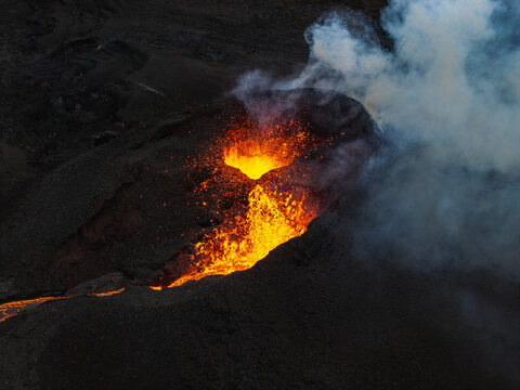 Aerial view of glowing lava cascading down the dark volcanic slopes amidst billowing smoke, Grindavik, Iceland.