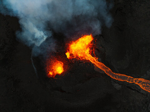 Aerial view of molten lava flowing like liquid fire against the stark black landscape, with plumes of smoke rising into the sky, Grindavik, Iceland.