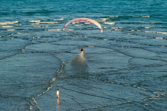 Aerial view of a parasailor gliding over shallow turquoise waters, with a lone figure standing on the shore, Jupiter, Florida, United States.