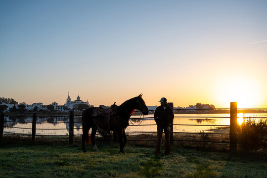 Cowboy Standing With Mustang Beside Lake At Sunrise