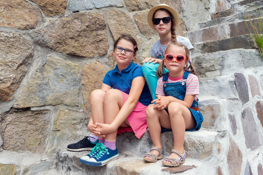 Three cheerful young girls in colorful summer outfits sit playfully on rustic stone outdoor stairs, smiling happily with sunglasses and hats, perfect for childhood friendship and vacation themes