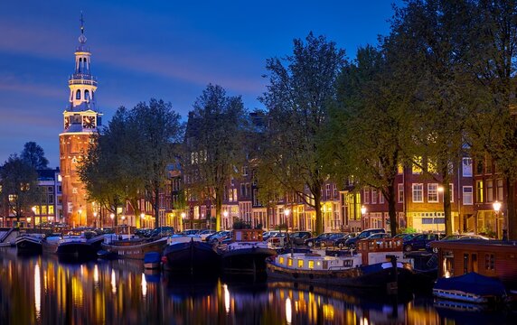Amsterdam, Netherlands. Houses with nighttime illumination over canals with water in Old town and view at the tower with clock. Quarter with traditional dutch houses. Night landscape. Blue hour..