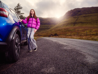 Fototapeta premium Young female driver in magenta color jacket is standing by a blue car, stunning nature scenery in the background with mountains and fields. Horse shoe drive, county Sligo, Ireland.