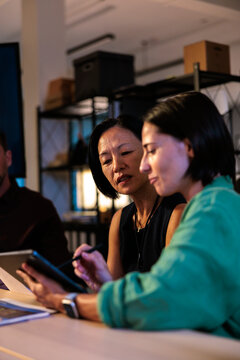 Two women collaborate on a tablet in a dim office.