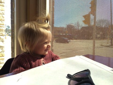 Smiling toddler in sweater sitting by sunny window at restaurant