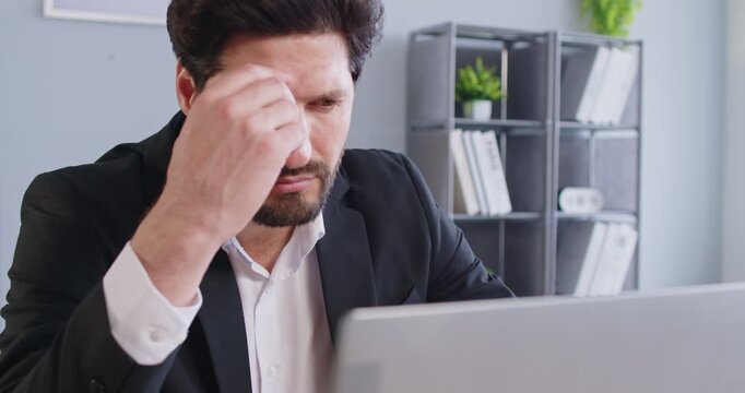 Worried businessman in suit sitting at desk in office and looking at laptop screen. Thoughtful male company employee trying to find solution while thinking about difficult work task during workday.