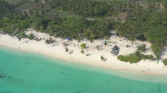 Aerial view of a relaxing beach on Thoddoo island with tourists swimming in clear turquoise water, sun umbrellas, green palm trees, and an old boat resting on the white sand.