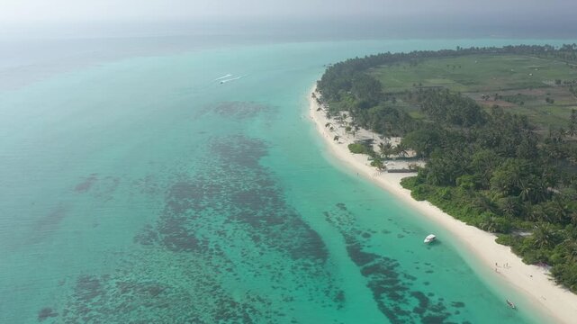 High altitude drone view of Thoddoo island featuring agricultural fields, a long white beach, vast coral reefs in turquoise water, and a speedboat sailing in the distance.