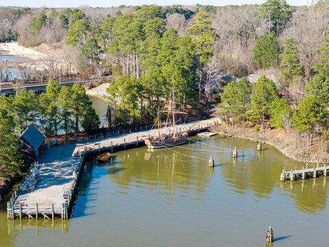 Colonial-era merchant ship replica, the Godspeed, docked at the wooden pier of the Jamestown Settlement living history museum on the James River. Aerial view.