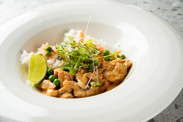 A bowl of creamy chicken curry served with white rice, garnished with green peas, fresh microgreens, and a wedge of lime on a light gray stone surface