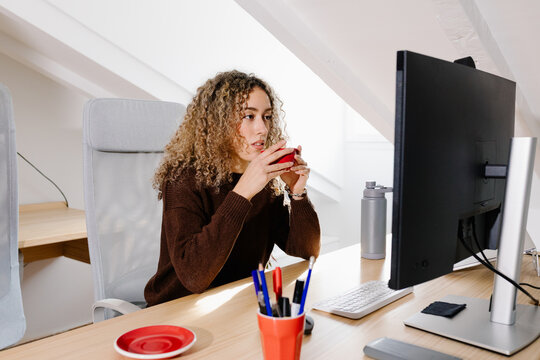 Self-employed woman participating in a video call and sipping coffee