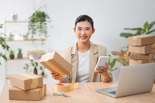 Asian woman preparing packages for shipment, using phone and laptop