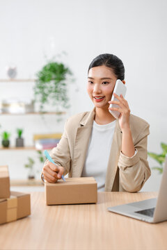 Asian woman on phone preparing package for shipping in home office