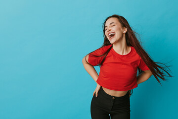 Young woman laughing with eyes closed standing against blue background wearing red shirt and black...