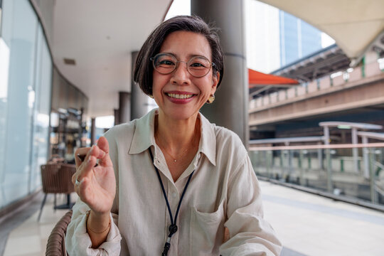 Smiling woman with glasses waves in an urban setting.