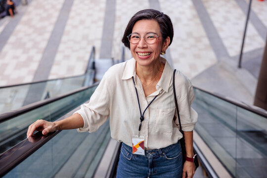 Smiling woman rides escalator, wearing glasses and name tag.
