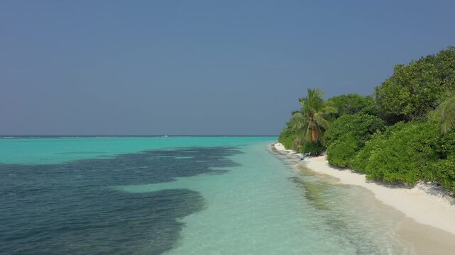 Low aerial view flying along the pristine Thoddoo island shoreline, showcasing dense green tropical vegetation, a narrow white sand beach, and crystal clear turquoise ocean water.