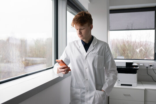 Researcher in White Coat Checks Phone by the Window 