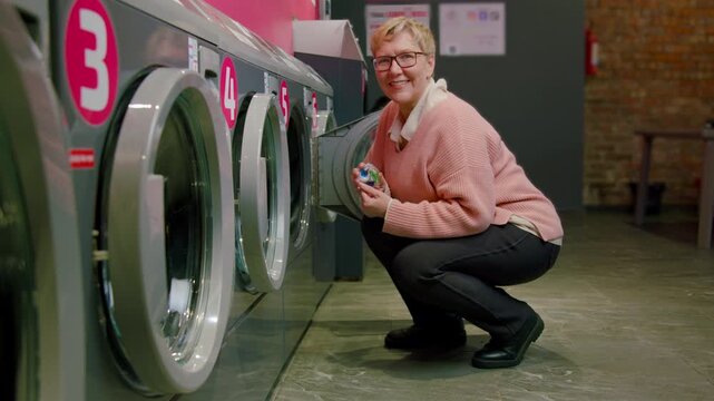 Caucasian woman in pink sweater squats near washing machines in a laundromat, holding laundry detergent while smiling in a bright, modern interior space