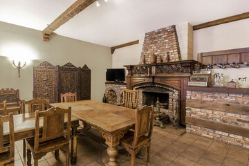Fototapeta premium A dining room features a large wooden table surrounded by chairs. A brick fireplace stands on one side, and a TV is mounted on the wall. The room is warm and inviting