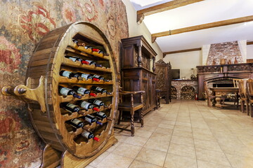 Fototapeta premium A wooden wine rack holds several bottles in a cozy room. The space features wooden furniture, a fireplace, and a decorative wall. The warm lighting creates a welcoming feel in the evening