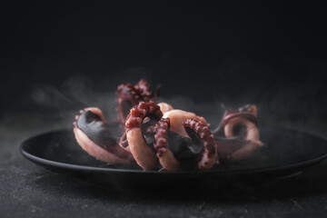 Hot prepared squid or boiled octopus with tentacles wiggling on a plate with steam coming out against black background. Seafood background for a restaurant menu © alexanderon