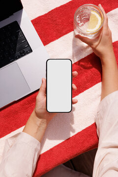 Person holding smartphone and drink by laptop on red towel