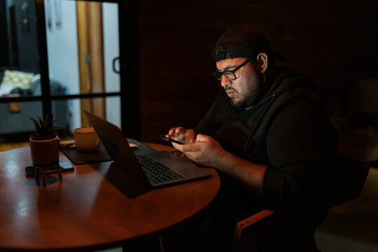 Man Checking Mobile Phone While Working On Laptop At Night