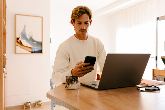 Young man multitasking in a modern room, using a laptop and smartphone