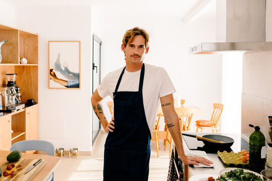 Modern man looking at the camera, posing in an apron at his kitchen