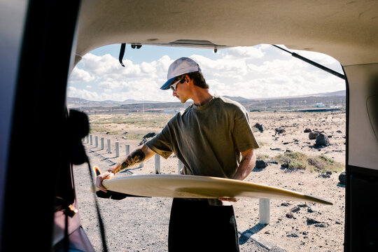 A young surfer pulling his surfboard out of the van&rsquo;s trunk