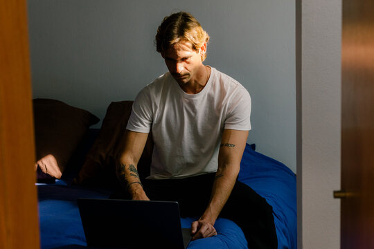 Young man working remotely on a laptop, seated on the edge of a bed