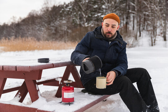 Hiker pouring hot coffee from camping pot on picnic bench in winter