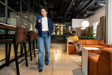 Fototapeta premium Young woman holding smartphone reading message walking in coworking space