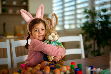 Little girl wearing bunny ears hugging a plush rabbit with decorated eggs on table