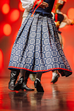 Latvian folk dance featuring traditional blue patterned skirt and black shoes, vibrant red background highlighting cultural attire and movement